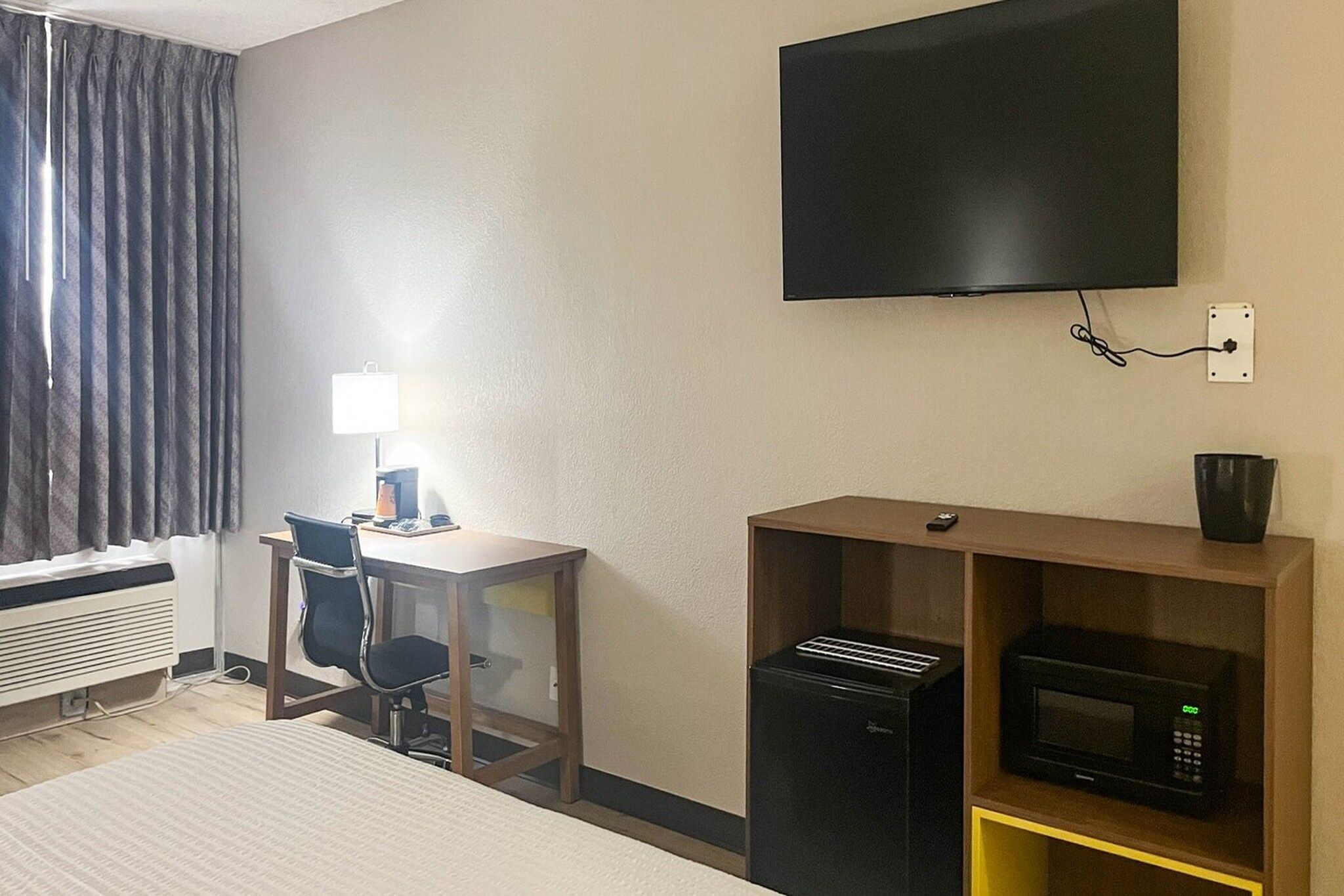 Ett hotellrum med en säng, ett skrivbord, en stol, en tv, en mikrovågsugn och en kaffemaskin.