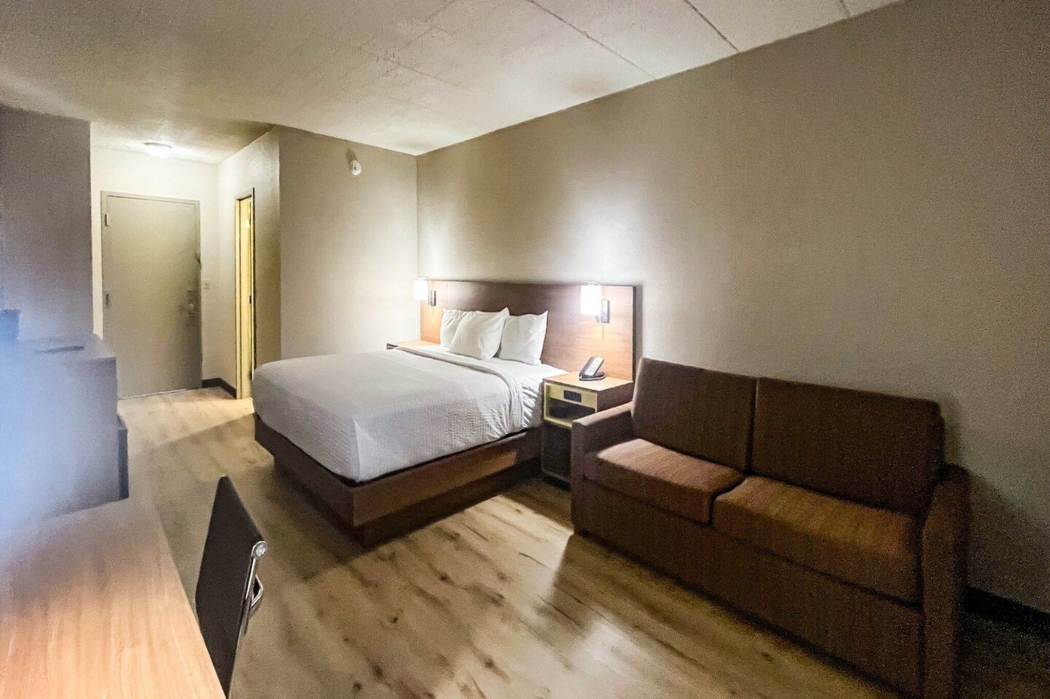 A hotel room with a bed, a bedside table with a phone, a sofa, and a wooden floor.