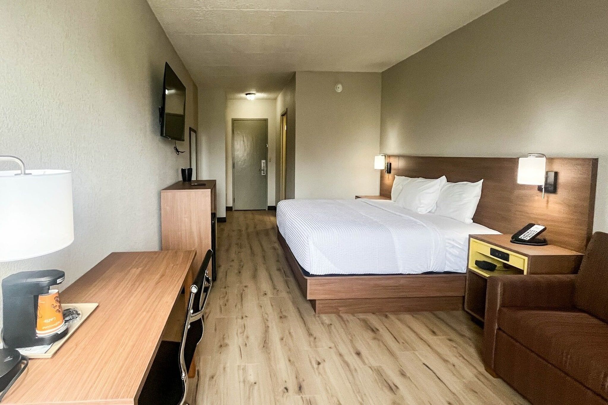 A hotel room with a bed, bedside tables, a desk with a lamp, a TV, and a door leading to a hallway.