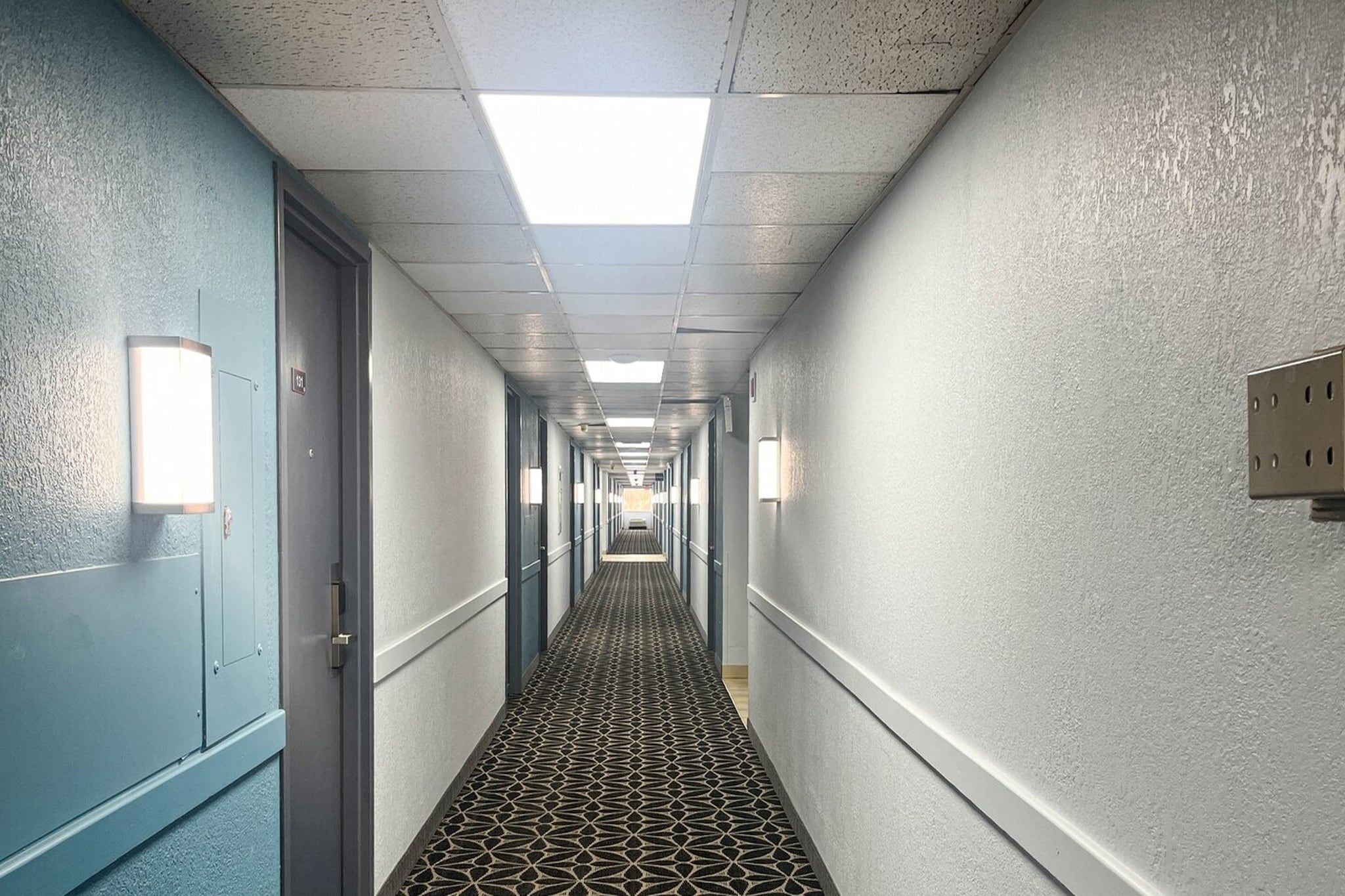 Um corredor de hotel comprido e estreito, com portas em ambos os lados, carpete estampado e luminárias fixadas na parede.