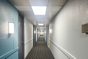 Un largo y estrecho pasillo de hotel con puertas a ambos lados, alfombra estampada y luminarias fijadas a la pared.