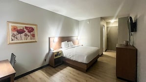 Quarto de hotel com cama, mesa de cabeceira, cadeira, escrivaninha e um quadro na parede.