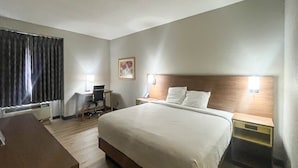 Quarto de hotel com uma cama grande, uma escrivaninha com cadeira, uma mesinha e uma mesa de cabeceira com luminária.