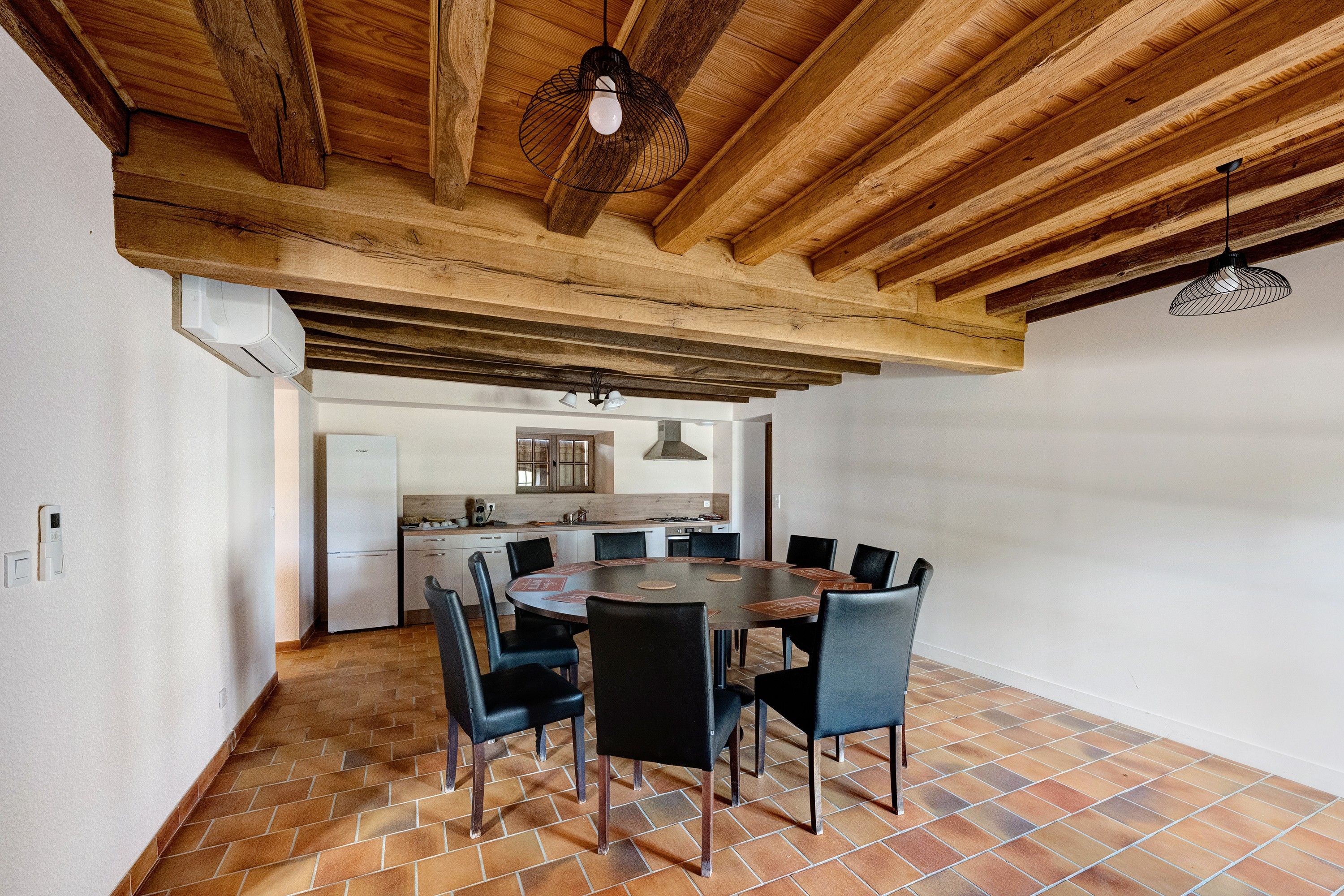 A spacious dining area with a round table and chairs, a kitchen in the background, and a wooden ceiling with exposed beams.