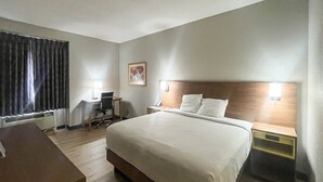 Quarto de hotel com uma cama grande, uma escrivaninha com cadeira, uma mesinha e uma mesa de cabeceira com luminária.