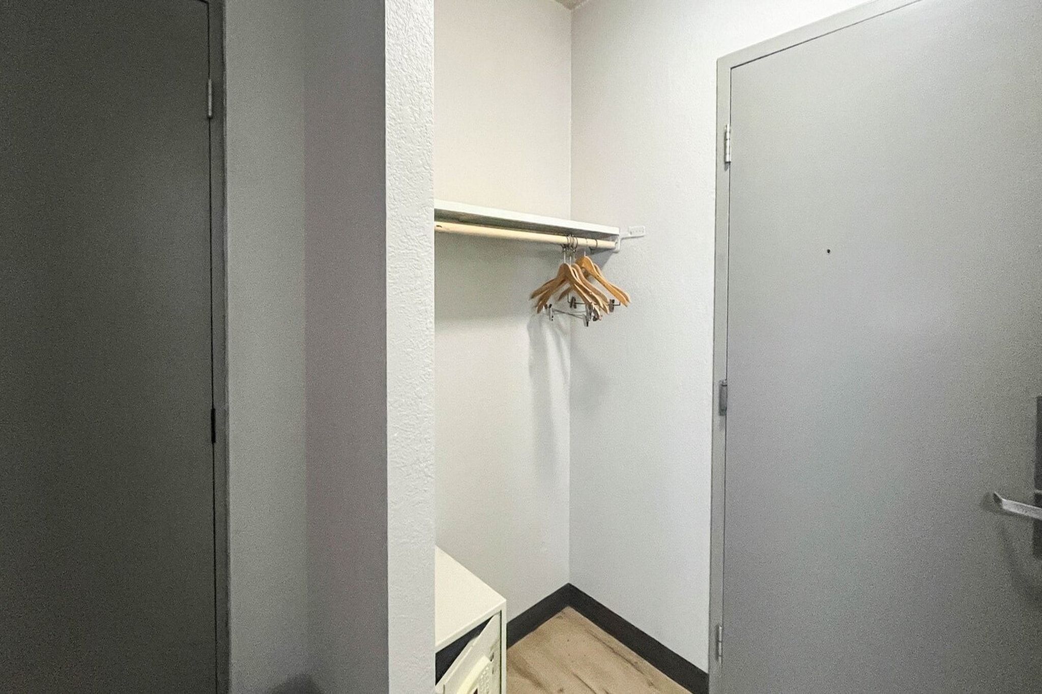 A small, narrow hallway with a white door on the right and a white wall with a wooden hanger rack on the left.
