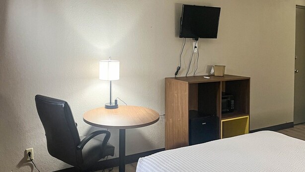 A hotel room with a bed, a desk with a chair, a lamp, a television mounted on the wall, and a small cabinet.
