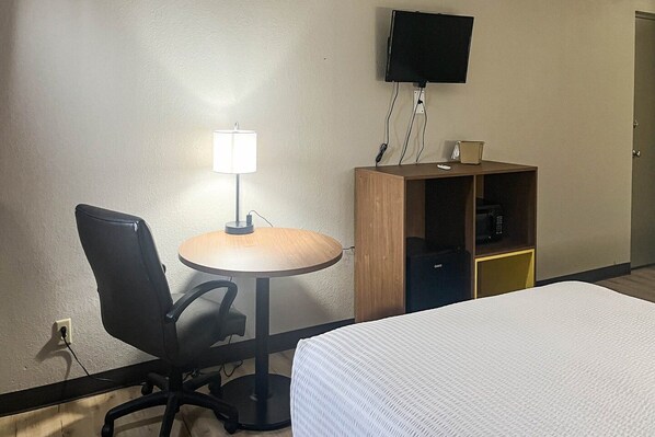 Habitación de hotel con cama, escritorio con silla, lámpara, televisor montado en la pared y un mueble pequeño.