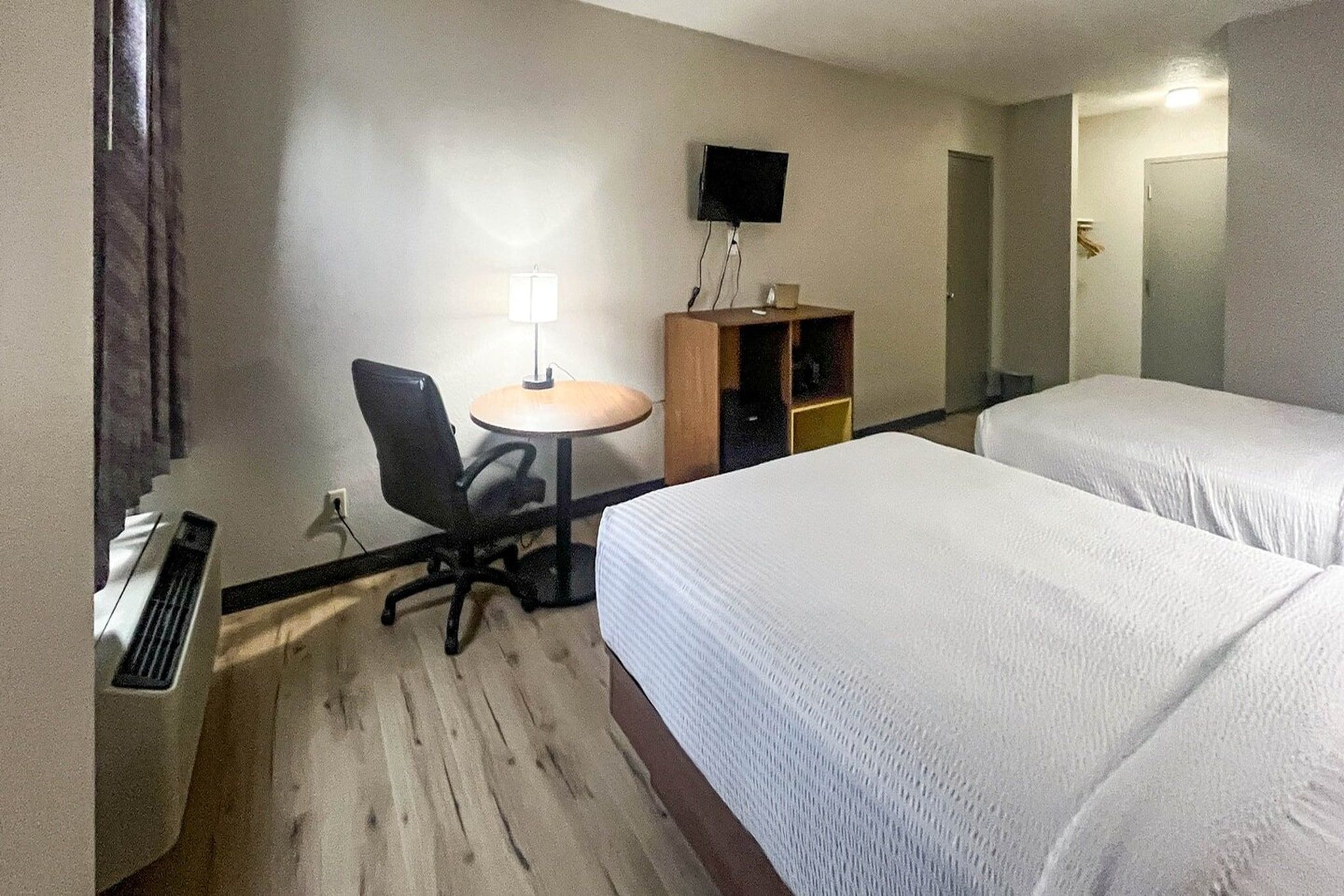 A hotel room with a bed, a desk with a chair, a small table, a television mounted on the wall, and a wardrobe.