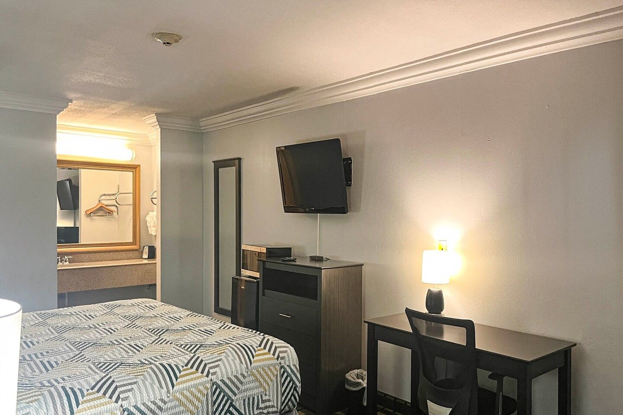 A hotel room with a bed, a desk, a chair, a television mounted on the wall, and a mirror above the mirror.