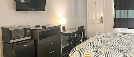 A hotel room with a bed, a desk with a lamp, a television, and a minibar.