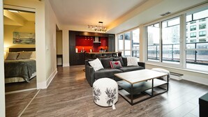 Una sala moderna con sofá, reposapiés y mesa de centro, una cocina con armarios rojos y ventanas amplias que ofrecen vistas a la ciudad.