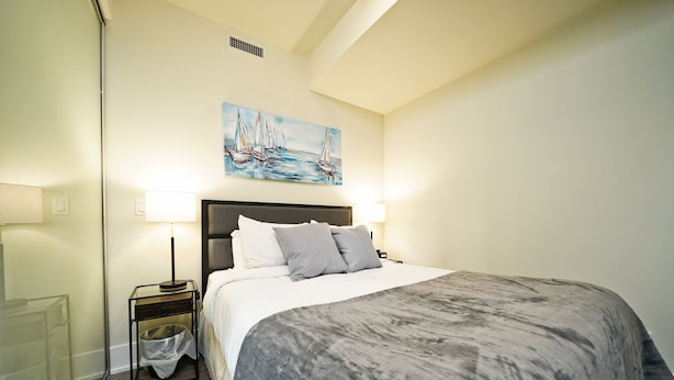 Un dormitorio con cama, lámparas de noche, un cuadro y una mesita de noche con un cubo de basura.