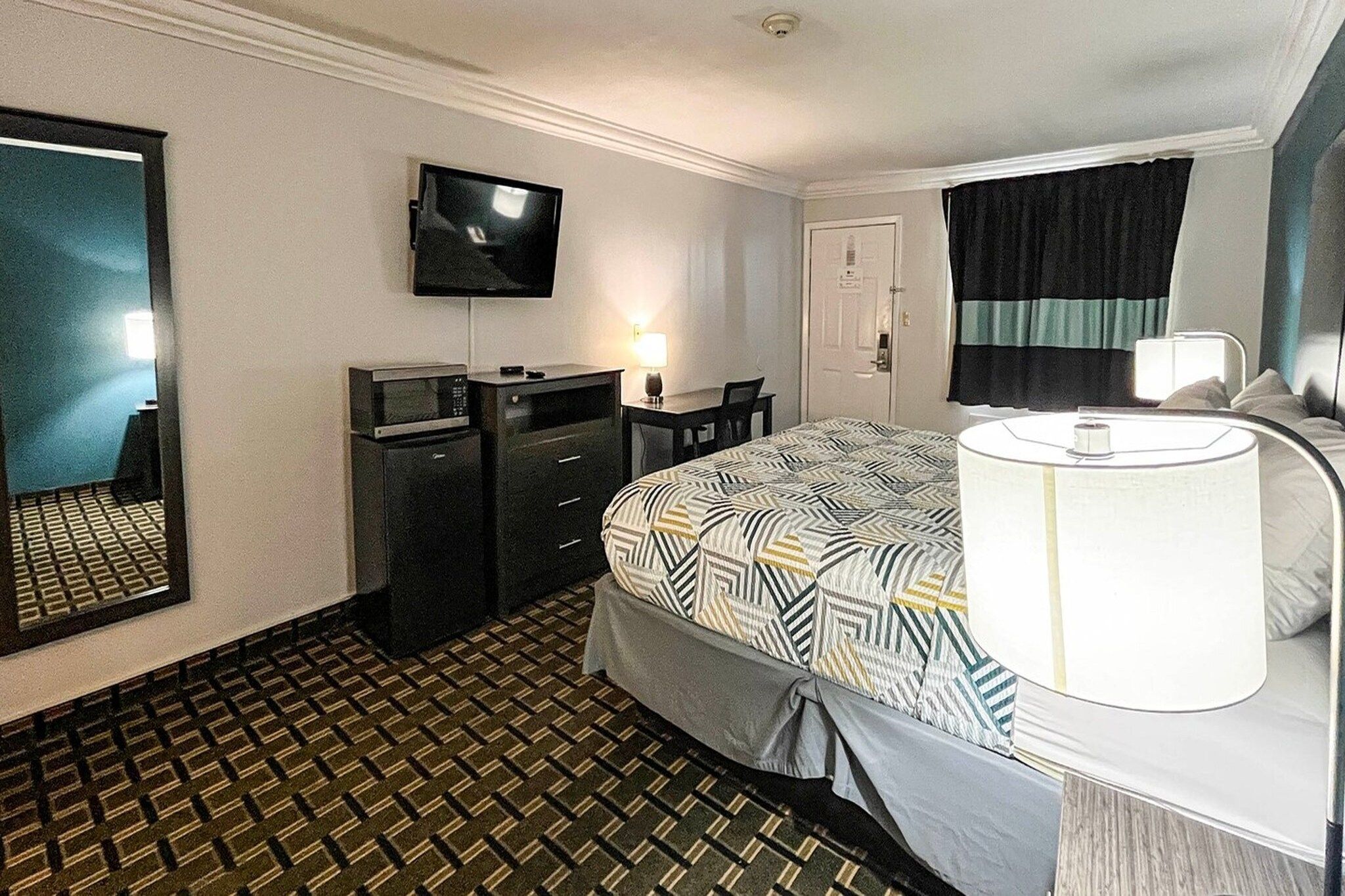 Quarto de hotel com cama, escrivaninha, cadeira, televisão, luminária de mesa e porta.