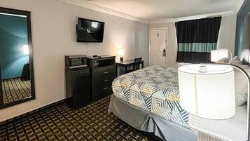 Quarto de hotel com cama, escrivaninha, cadeira, televisão, luminária de mesa e porta.