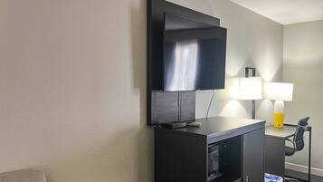 Et hotelværelse med et fladskærms-TV monteret på et skab, et skrivebord med lampe og en skraldespand.