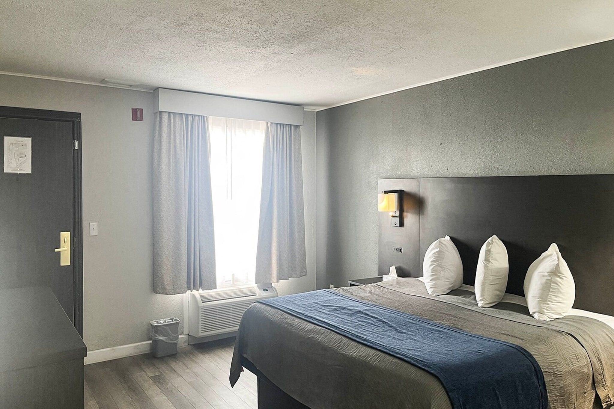 A hotel room with a bed, a bedside table, a window with curtains, a wall-mounted light, and a radiator.