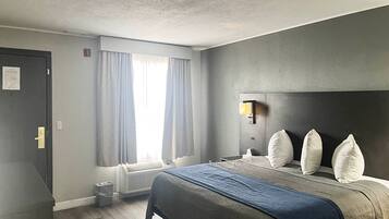 A hotel room with a bed, a bedside table, a window with curtains, a wall-mounted light, and a radiator.