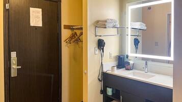 A hotel bathroom with a dark wooden door, a mirror with a lighted frame, a hairdryer, and a sink with a towel rack above it.