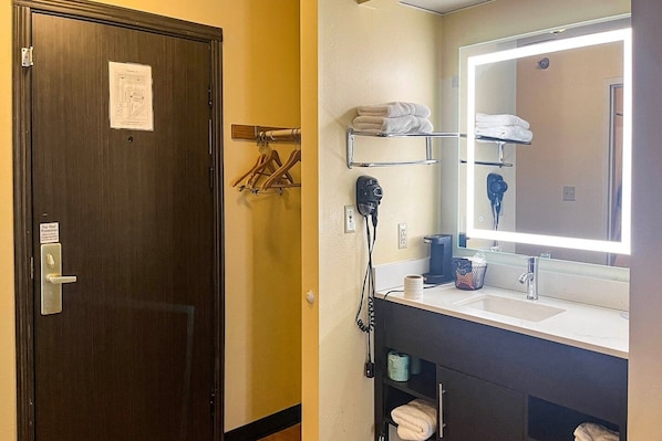 A hotel bathroom with a dark wooden door, a mirror with a lighted frame, a hairdryer, and a sink with a towel rack above it.