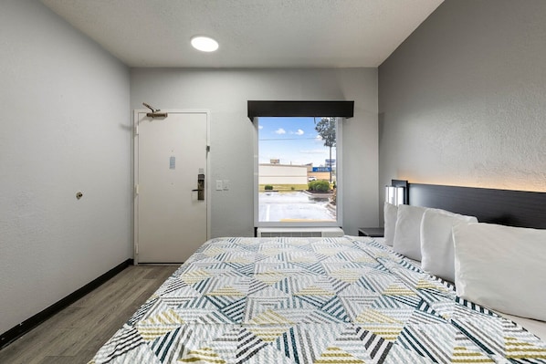 A hotel room with a bed, a door, a view of the outside, and a nightstand with a lamp.