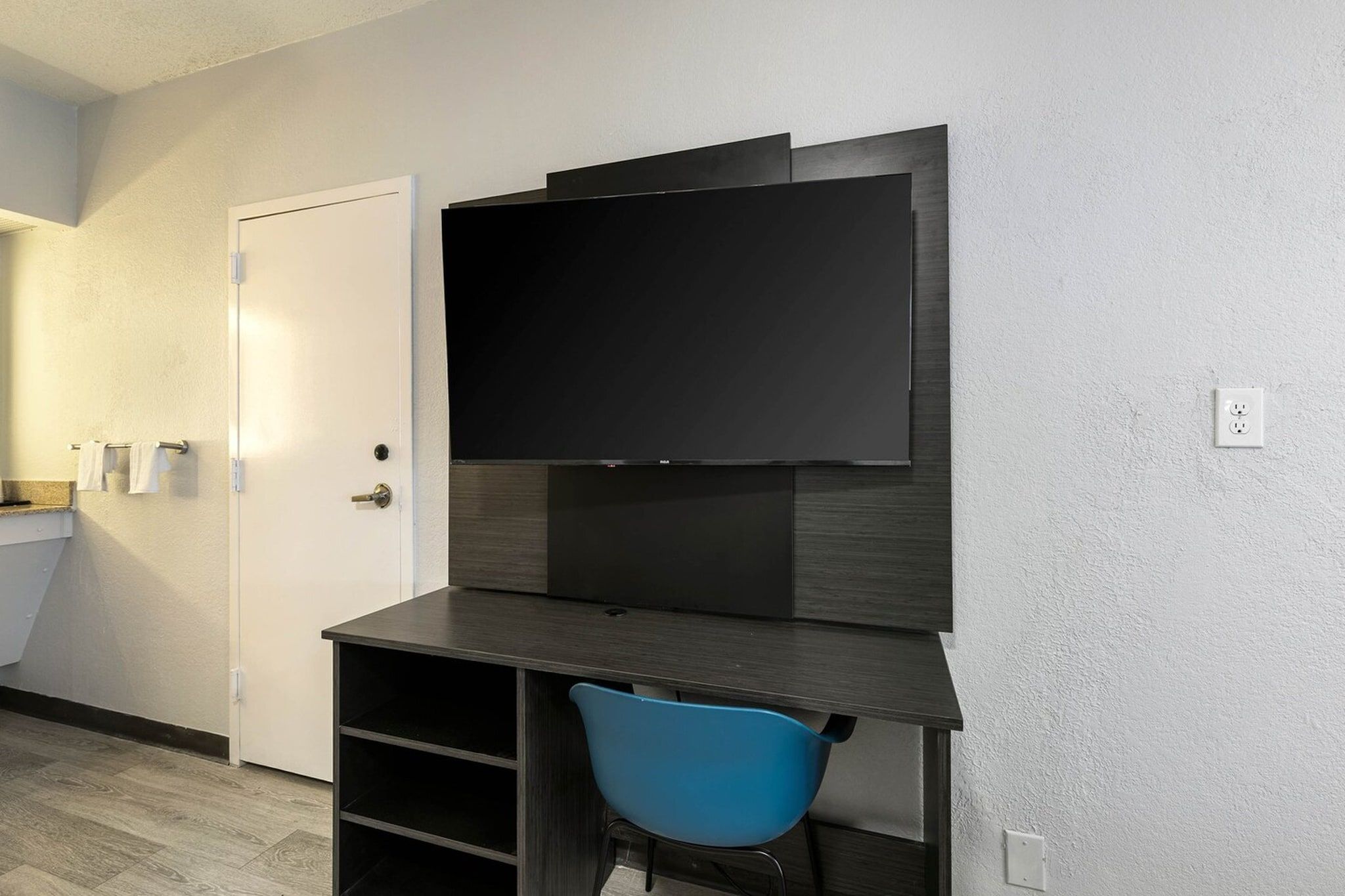 A small room with a flat-screen TV mounted on a dark wooden desk, a blue chair, a white door, and a towel rack.