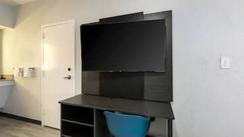 A small room with a flat-screen TV mounted on a dark wooden desk, a blue chair, a white door, and a towel rack.
