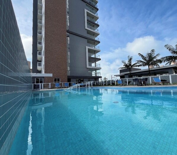 Una piscina amb una terrassa enrajolada, envoltada d'edificis moderns amb balcons i un cel serè.