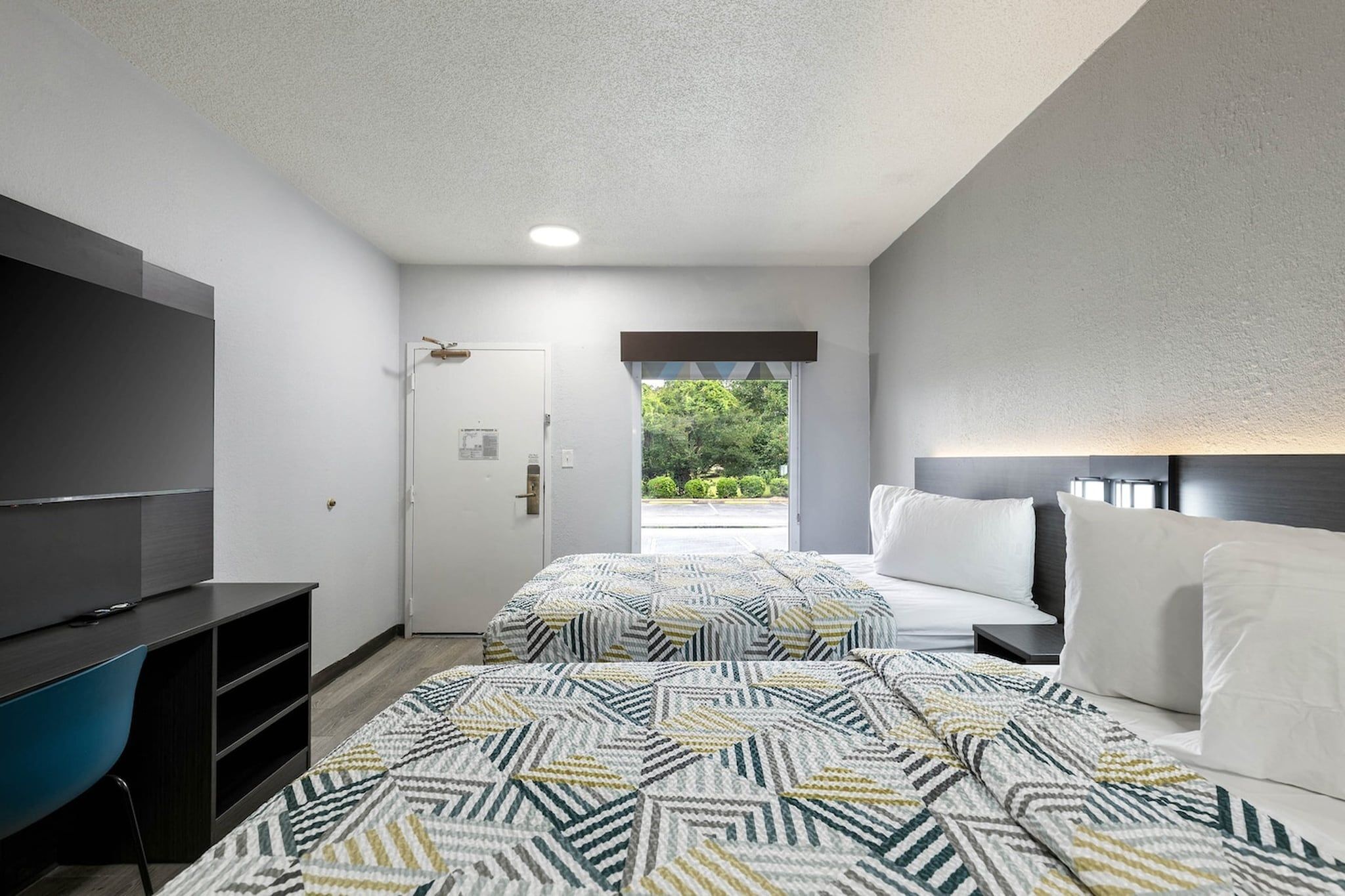 Quarto de hotel com televisão, cama com roupa de cama branca, tapete estampado e vista para o verde lá fora.