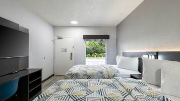 Quarto de hotel com televisão, cama com roupa de cama branca, tapete estampado e vista para o verde lá fora.