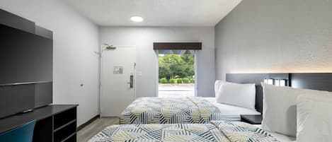 A hotel room with a TV, a bed with white bedding, a patterned rug, and a view of greenery outside.