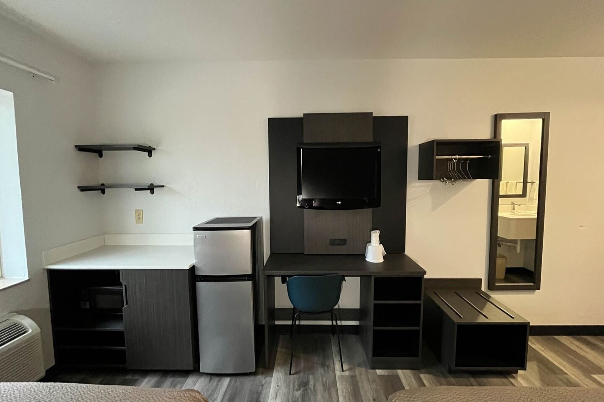 Una habitación compacta con kitchenette, televisión de pantalla plana, espejo y una silla.