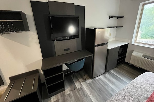 A compact hotel room with a kitchenette, a flat-screen TV mounted on the wall, a small desk with a chair, and a window providing a view of the outdoors.