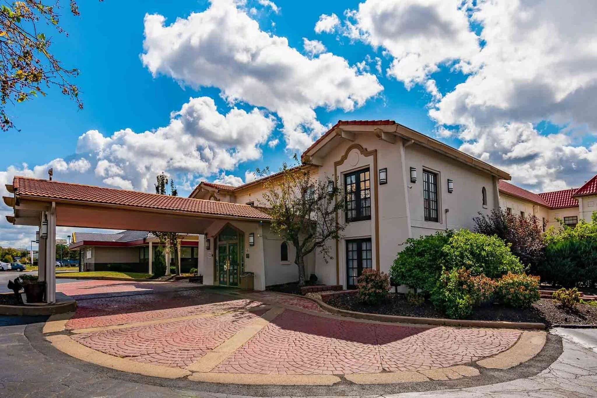 Um edifício com telhado de telhas vermelhas, entrada coberta e área ajardinada com calçada pavimentada.