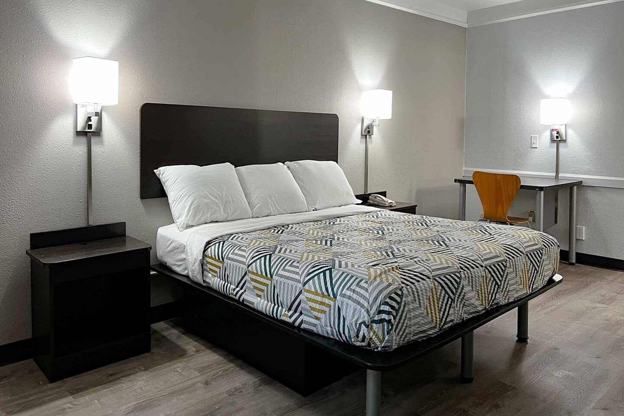 Quarto de hotel com cama, mesas de cabeceira, escrivaninha e duas luminárias fixadas na parede.
