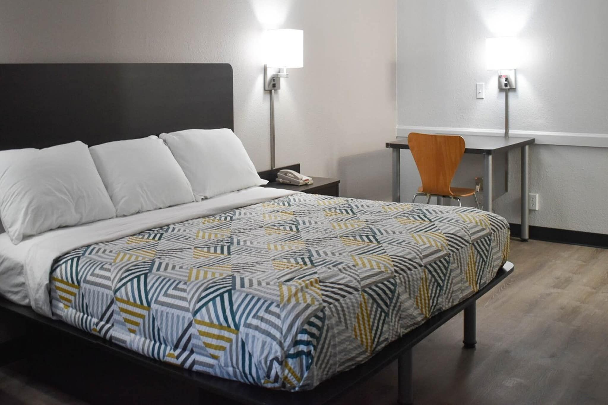 Une chambre d’hôtel comprenant un lit, une table de chevet, une chaise, un bureau et deux lampes fixées au mur.