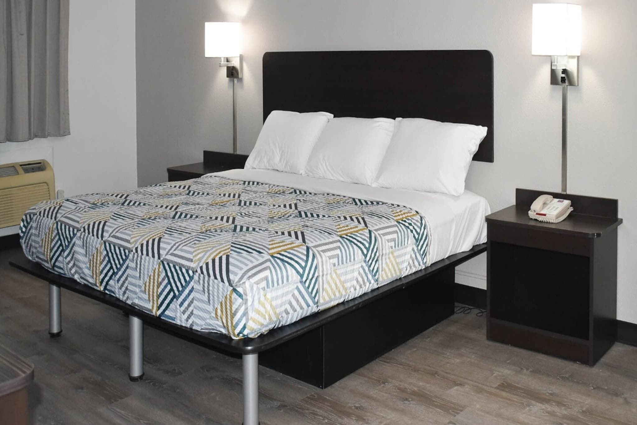 Quarto de hotel com cama, duas mesas de cabeceira, telefone e luminária na parede.