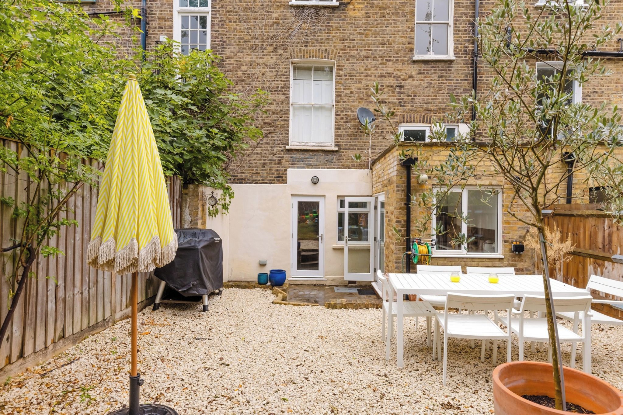 Un jardin avec une table et des chaises blanches, un parasol jaune et, au fond, une maison en briques.