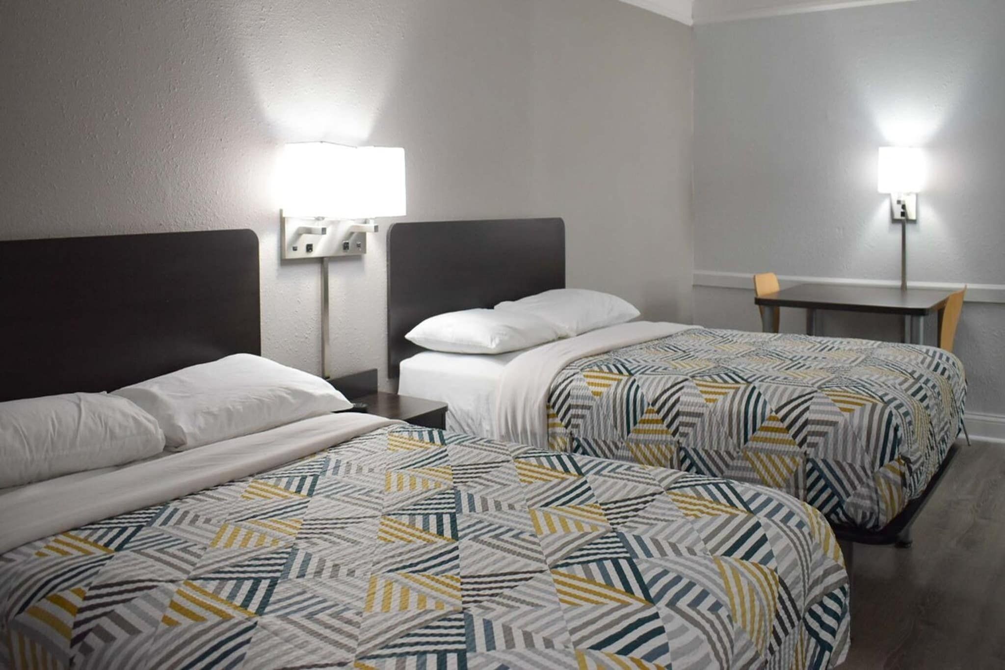 Une chambre d’hôtel avec deux lits, chacun recouvert d’une couvre-lit à motifs, une table de chevet avec une lampe et une petite table avec une chaise.