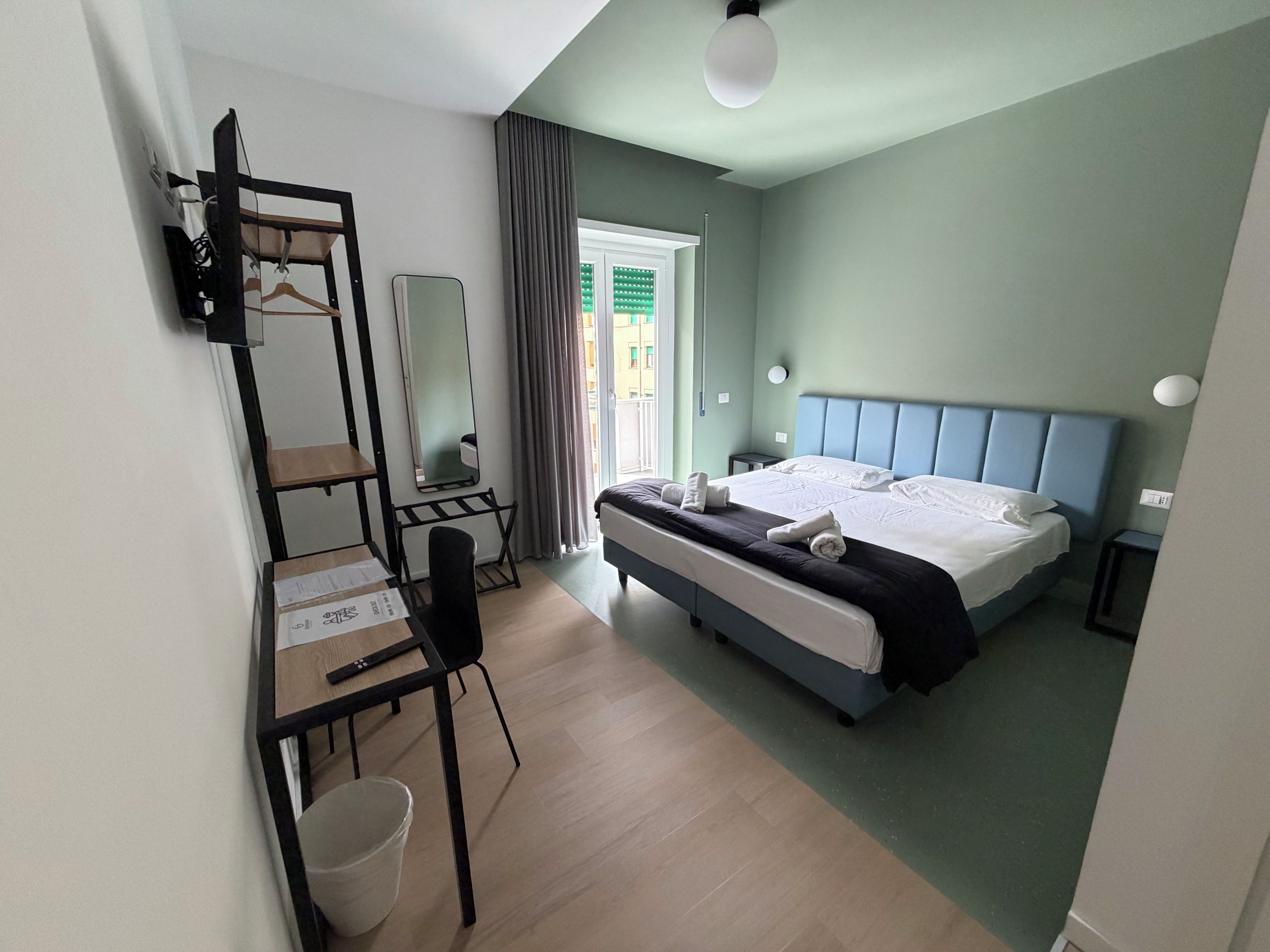 A hotel room with a bed, a desk, a chair, a mirror, and a window with curtains.