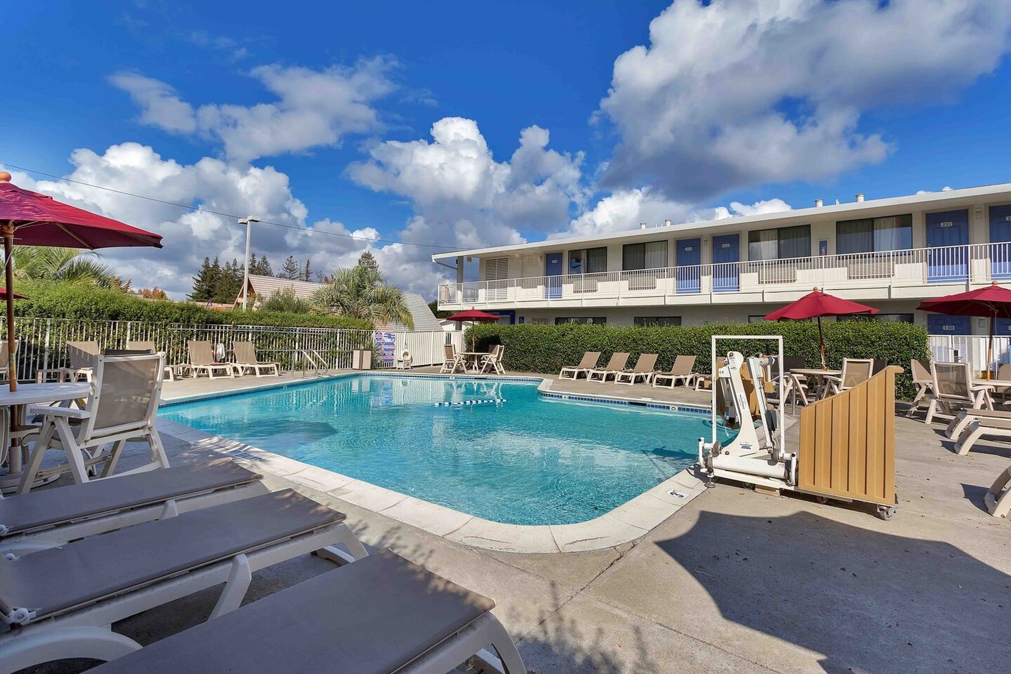 A swimming pool with lounge chairs and umbrellas, a multi-story building with balconies, and a clear sky.