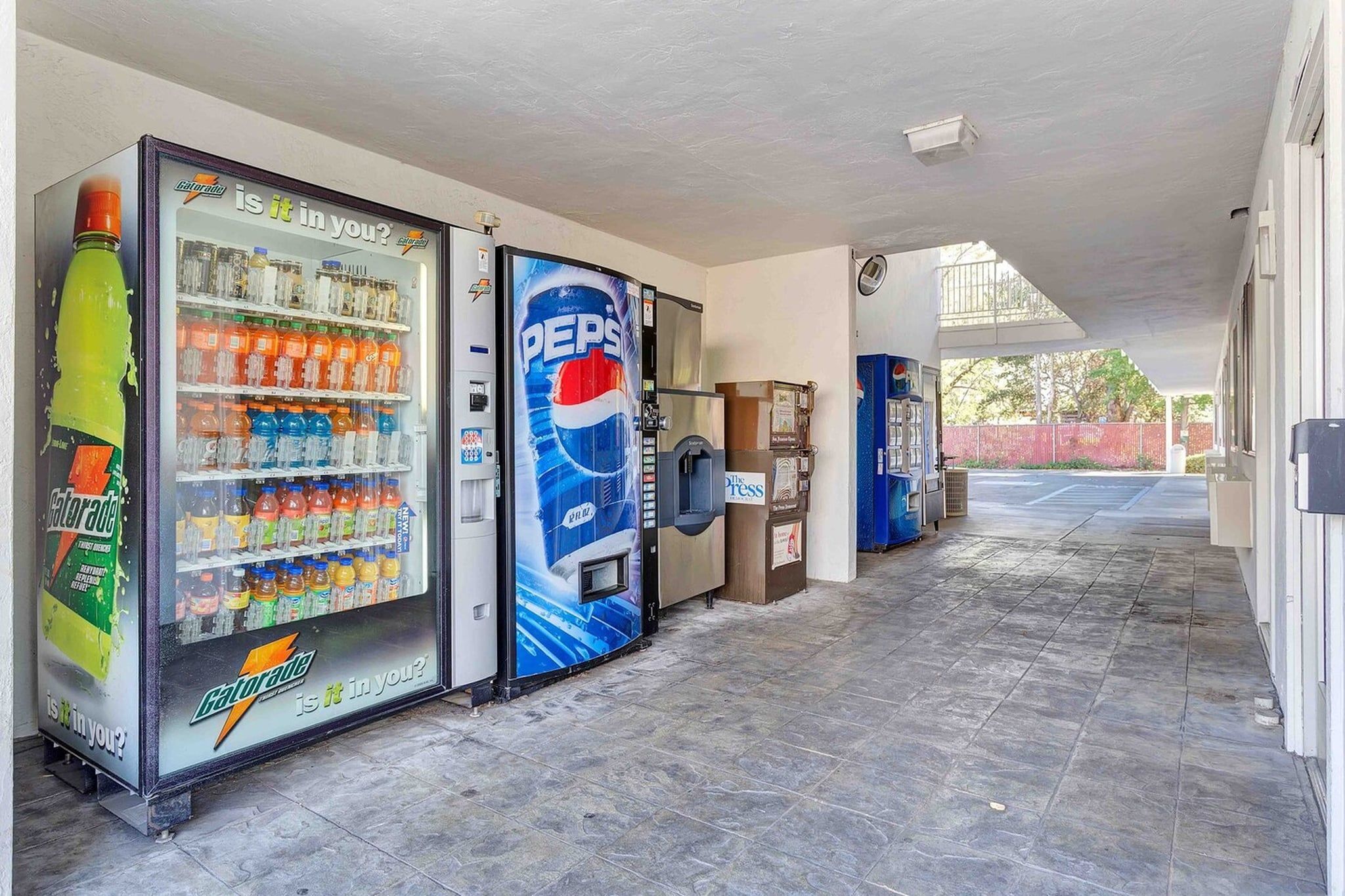 A corridor with vending machines selling beverages, a trash bin, and a covered area with a view of the outside.