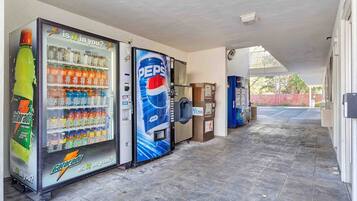 A corridor with vending machines selling beverages, a trash bin, and a covered area with a view of the outside.