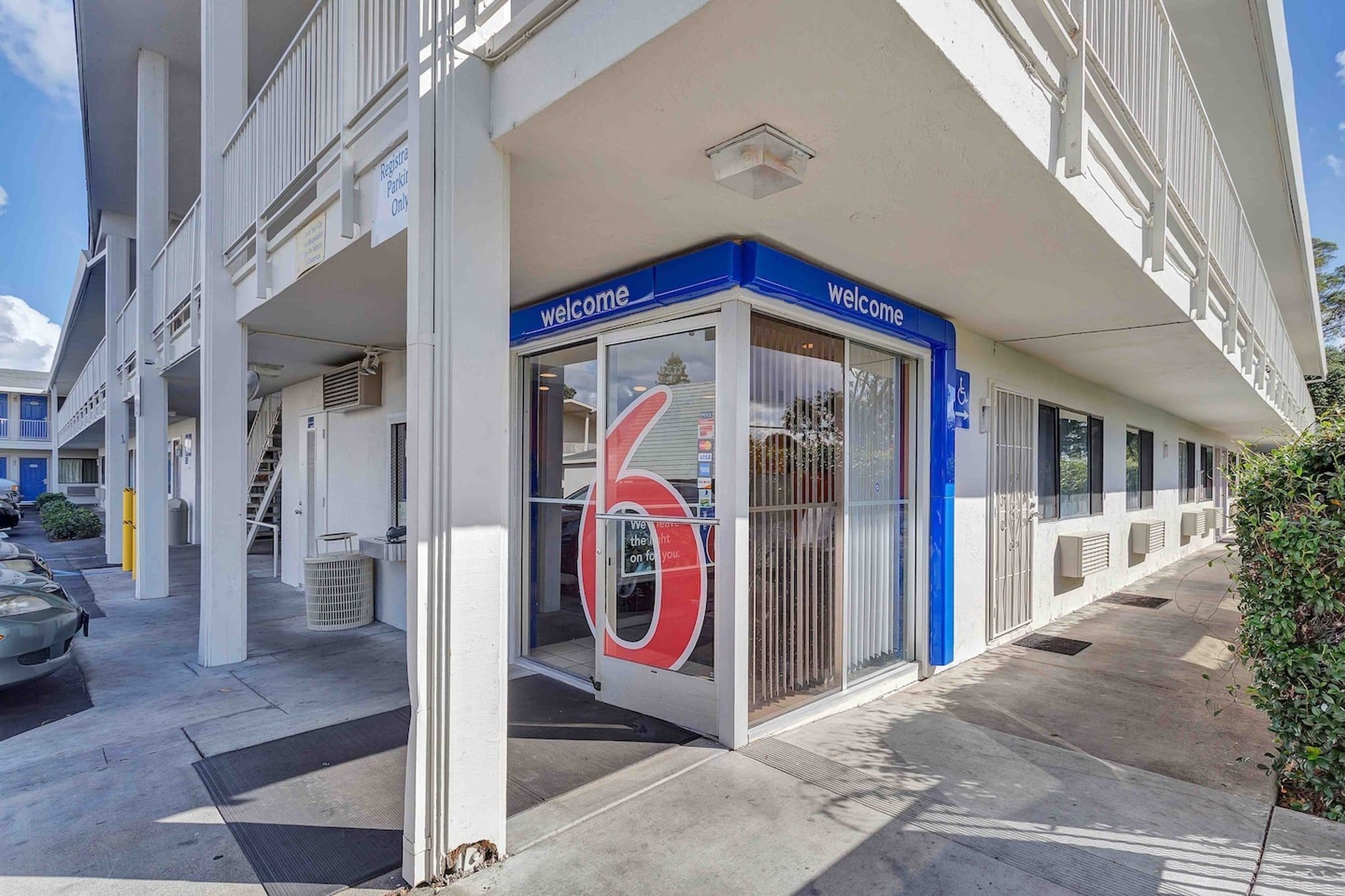 A building with a 'Welcome' sign, a large number '6' sign, and a parking area with a 'Register Parking Only' sign.