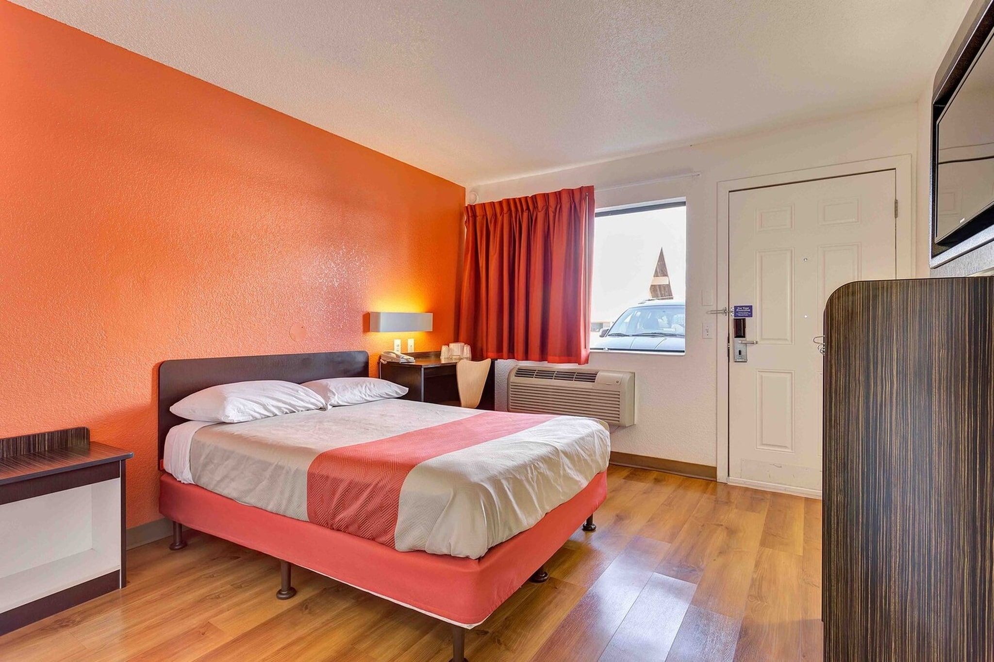 A hotel room with a bed, bedside table, lamp, window with curtains, and a door.