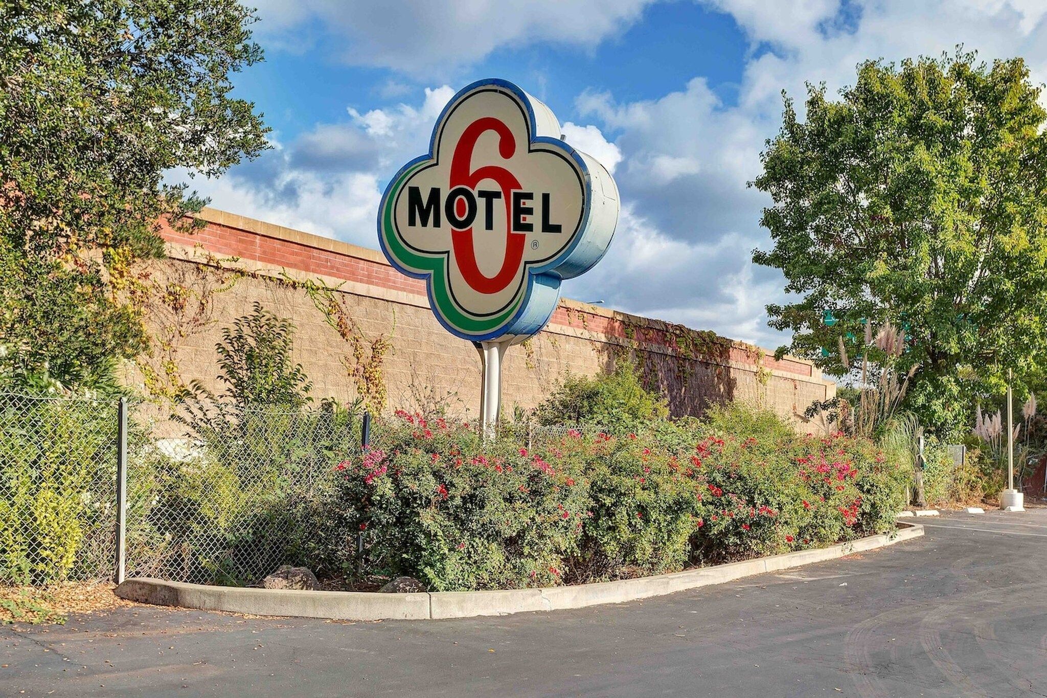 A motel sign with a number 6, surrounded by greenery and a fence.