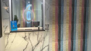 Uma área de recepção com piso de mármore, uma porta de vidro com o número '6' refletido e uma parede colorida listrada.