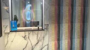 Uma área de recepção com piso de mármore, uma porta de vidro com o número '6' refletido e uma parede colorida listrada.
