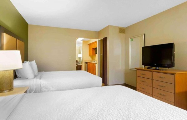 A hotel room with two beds, a television, a desk with a lamp, and a kitchenette.