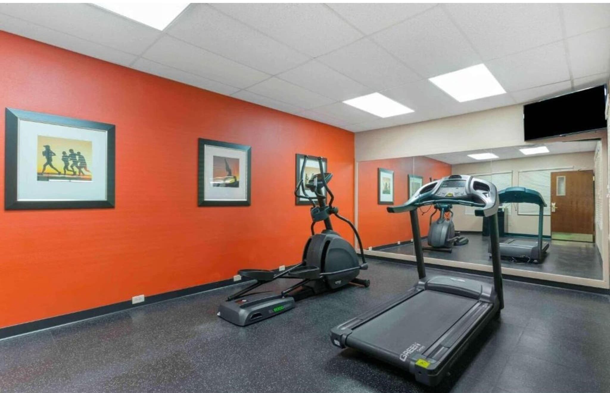 Ein Fitnessraum mit Trainingsgeräten, einer Spiegelwand und gerahmten Bildern an der orangefarbenen Wand.